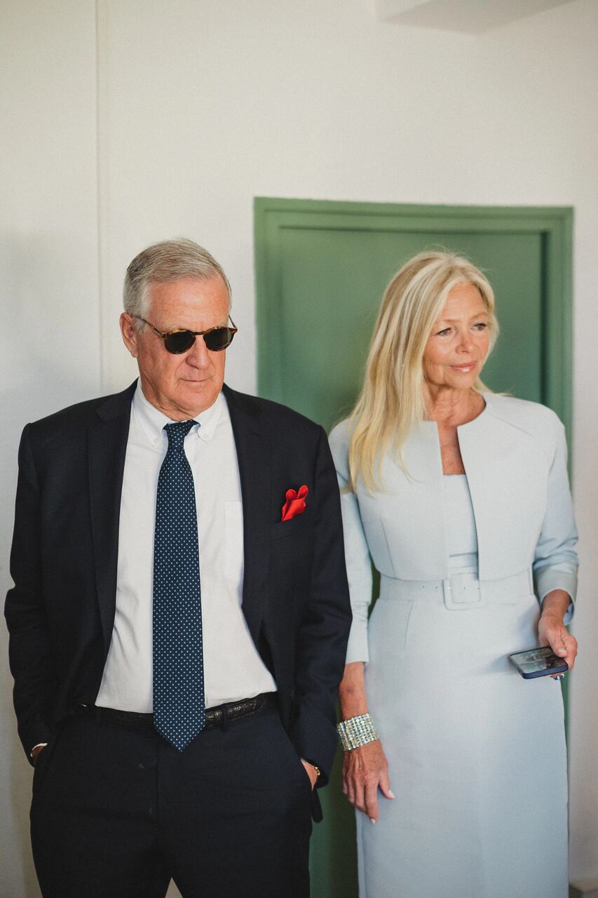 Casal elegante em ambiente interno; homem de terno preto com lenço vermelho no bolso e mulher usando vestido azul claro sofisticado.