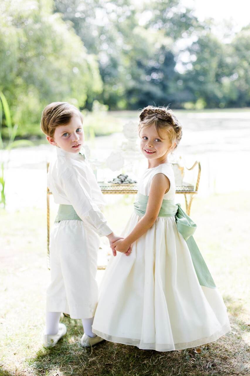 Um pajem e uma florista posam de mãos dadas em trajes clássicos com detalhes verdes, em um cenário à beira de um lago.