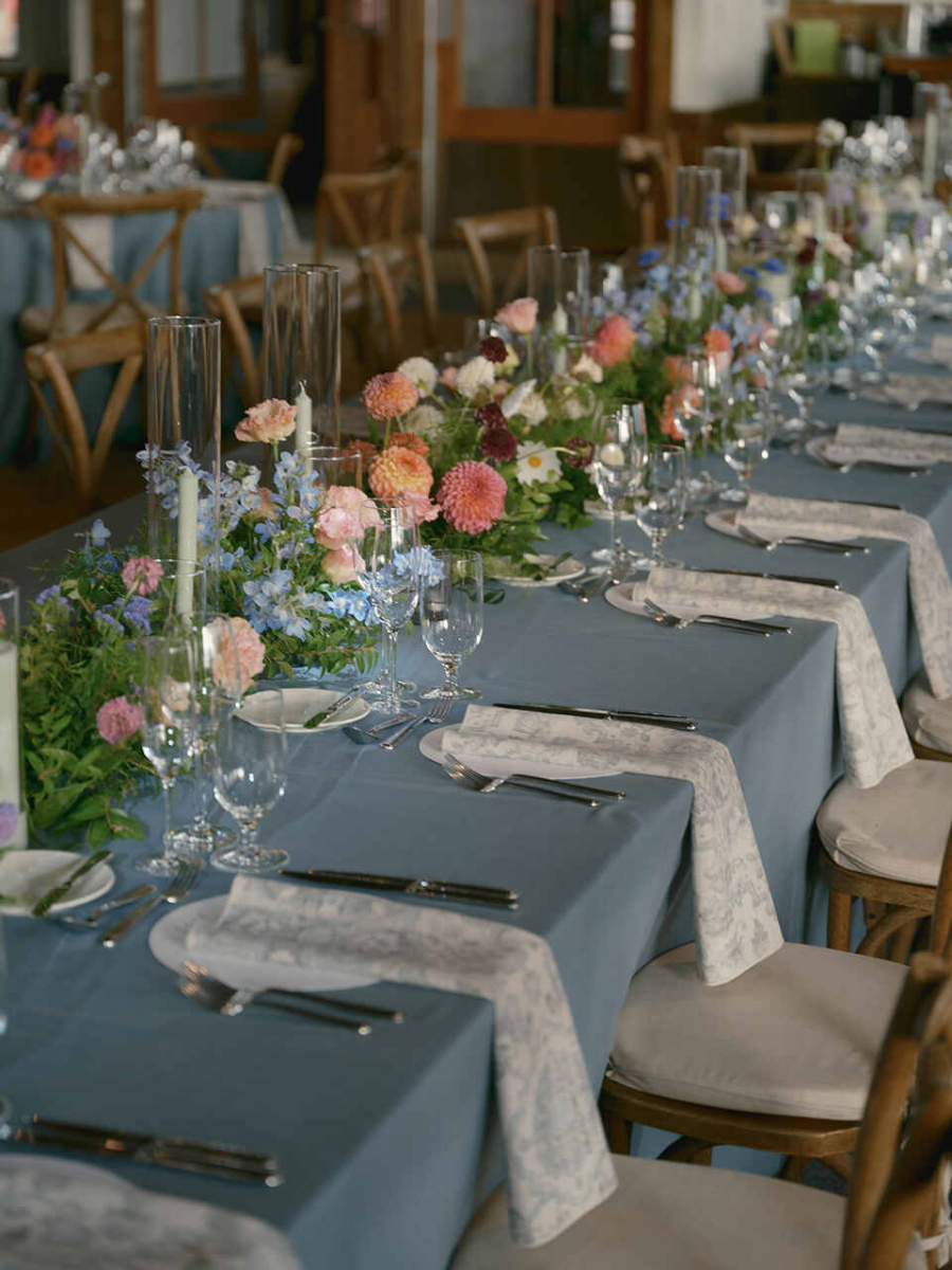 Uma mesa longa com toalha azul e delicados arranjos de flores pastel