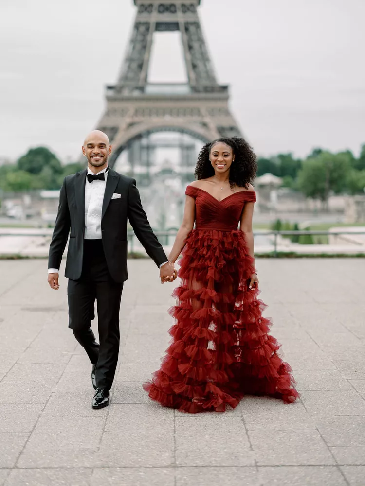 Casal posando em frente à Torre Eiffel; homem de smoking preto e mulher em vestido longo vermelho com camadas de tule.