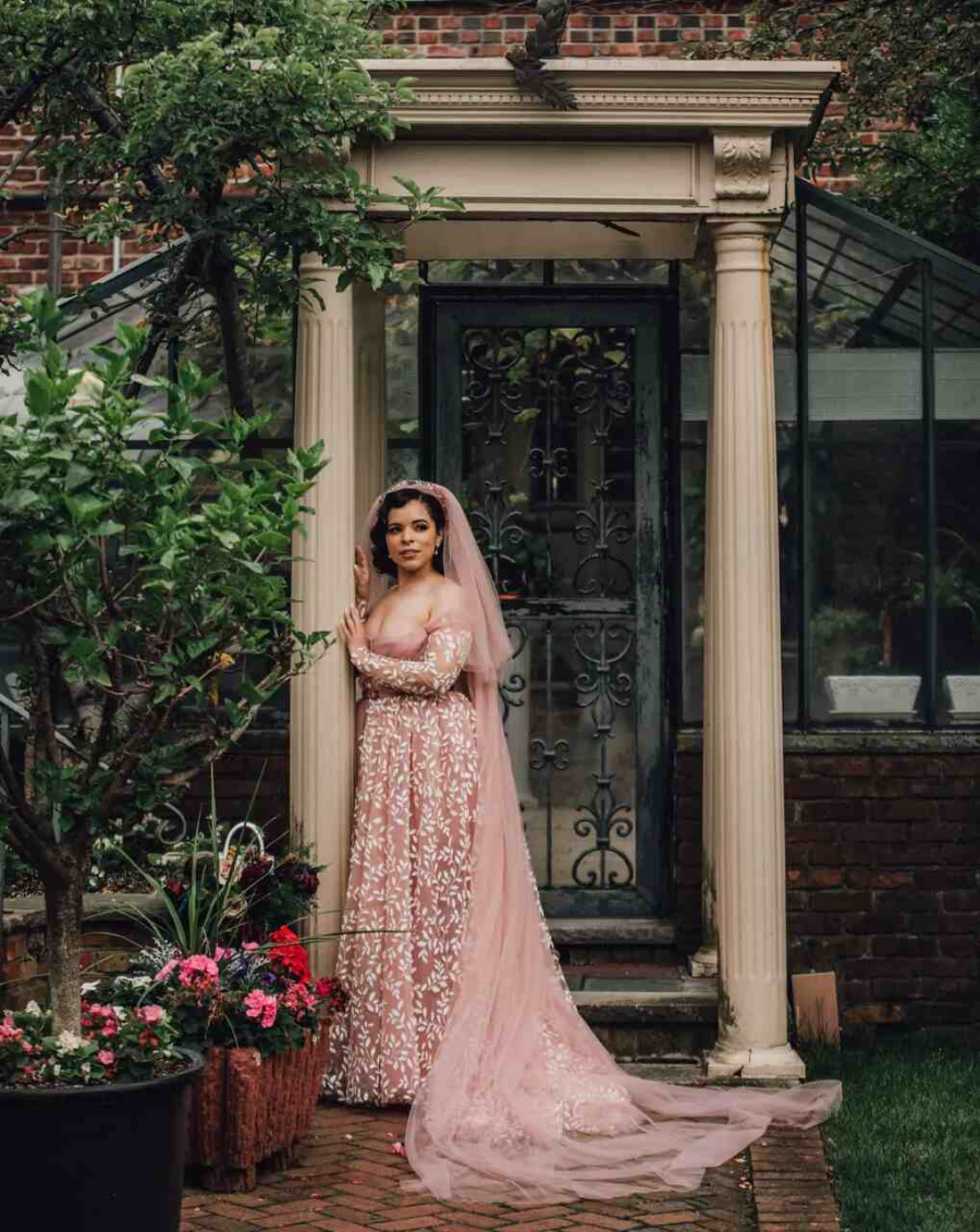 Noiva em um jardim elegante, com vestido rosa de renda detalhada e um véu longo. Ela está de pé próxima a uma estrutura clássica com pilares