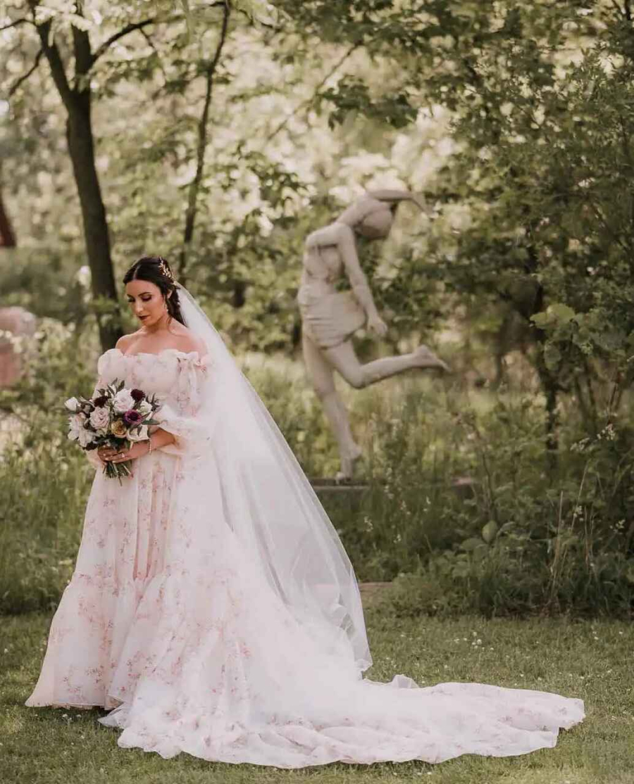 Noiva em um ambiente natural, usando um vestido floral em tons suaves com mangas bufantes e um véu longo. Ela segura um buquê elegante em tons de roxo e creme, enquanto uma estátua ao fundo complementa o cenário romântico e sereno.