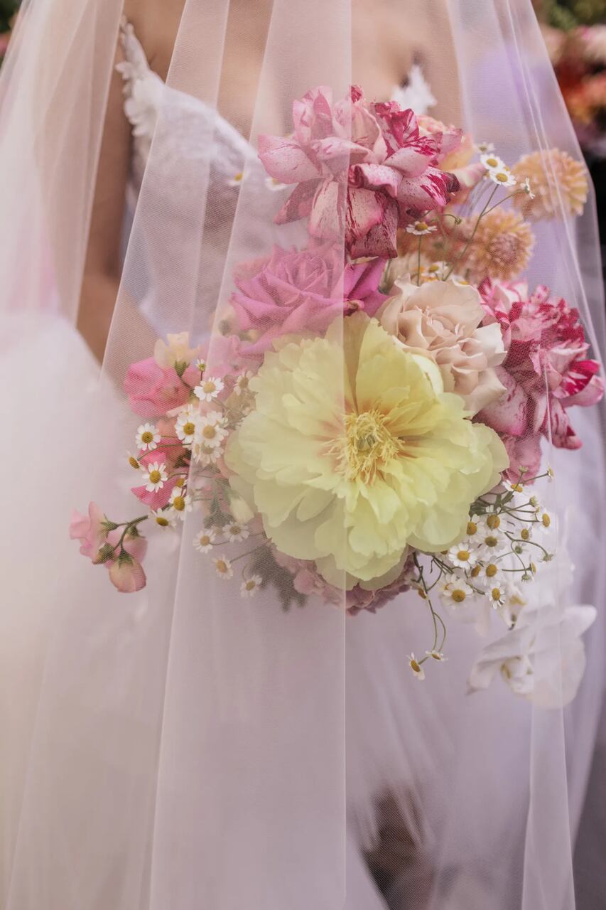 Uma noiva segurando um buquê exuberante com flores em tons de amarelo, rosa e branco, delicadamente emoldurado pelo véu translúcido.