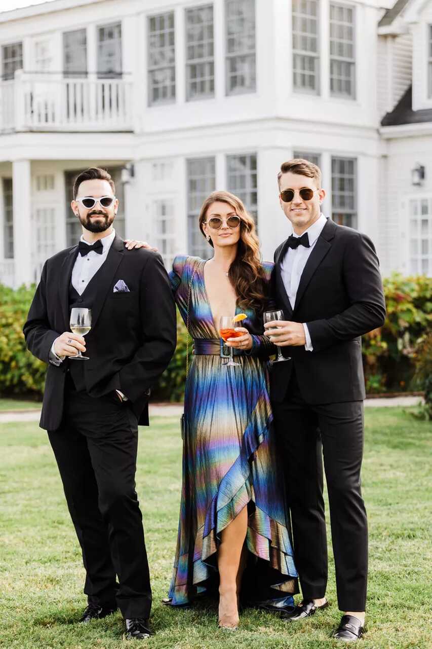 Grupo elegante em frente a uma mansão; dois homens de smoking e uma mulher com vestido metálico de listras coloridas, segurando taças de vinho.