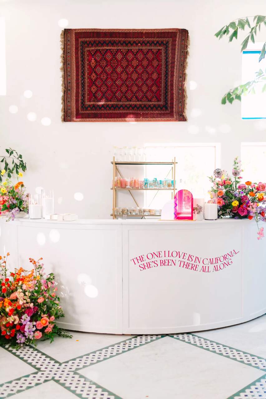 Mesa de bebidas decorada com flores coloridas e um painel de tapete artesanal ao fundo