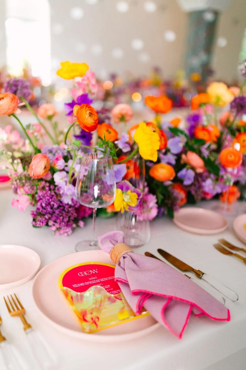 Detalhe de um prato decorado com flores vibrantes, taças de cristal e guardanapo rosa