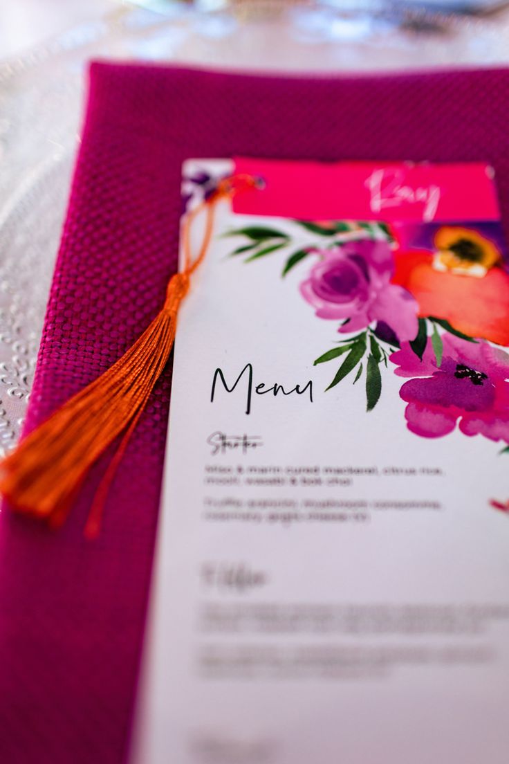 Menu de casamento com design floral colorido sobre guardanapo rosa vibrante, decorado com detalhe de tassel laranja