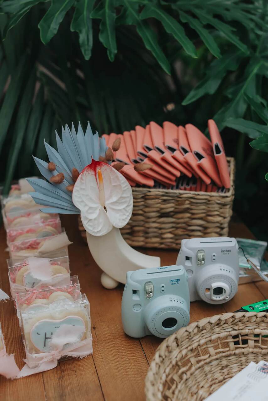 Um conjunto de câmeras Instax em tons pastéis dispostas sobre uma mesa de madeira, acompanhadas de biscoitos decorados e embalados individualmente. Ao fundo, leques coloridos em cestas e um arranjo tropical com folhas e flores vibrantes complementam o cenário.