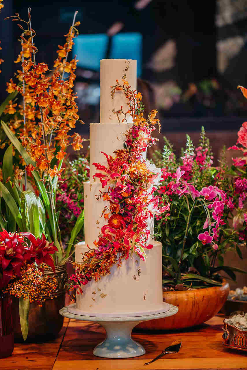 Bolo alto decorado com uma cascata de flores vibrantes, em uma mesa cercada por arranjos florais.