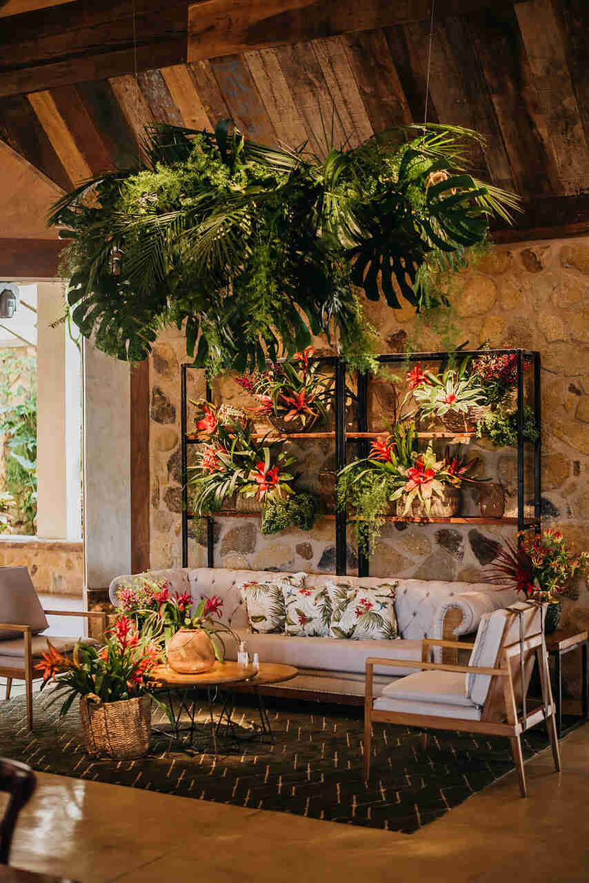 Lounge rústico com sofá floral, prateleiras decoradas com plantas tropicais e uma luminária suspensa de folhagens