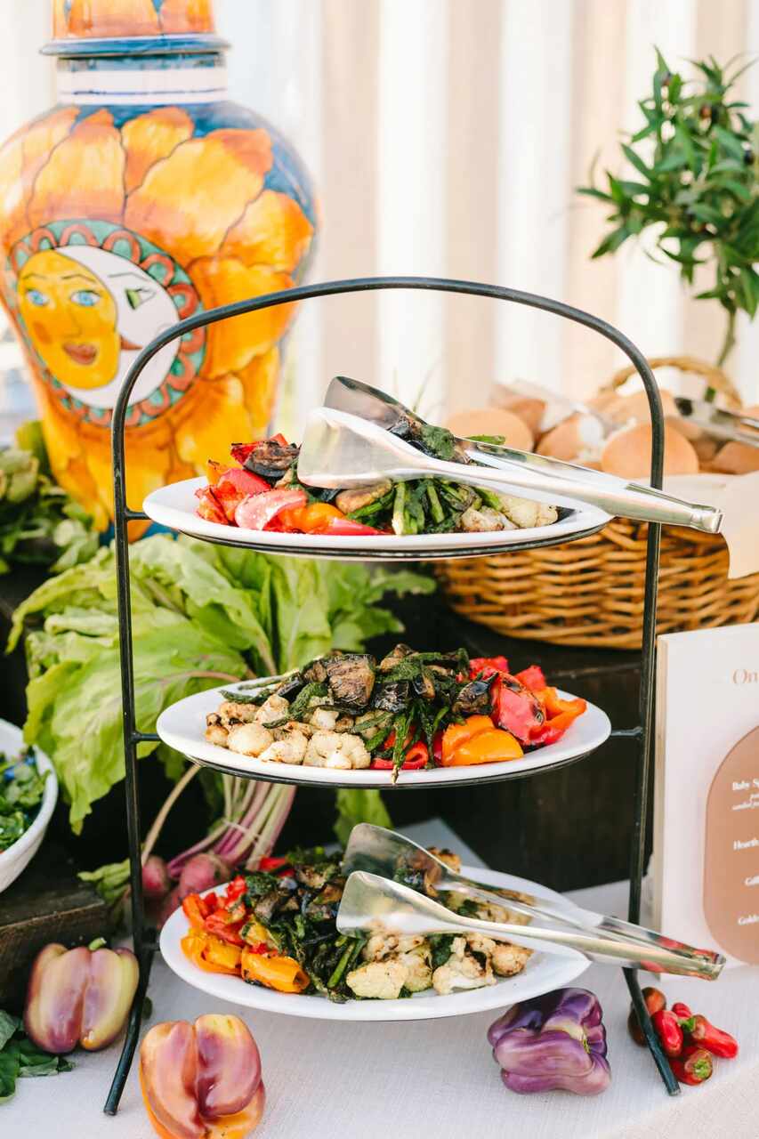 Estação de buffet com pratos de legumes grelhados dispostos em suporte de três níveis, ao lado de decoração colorida e temática