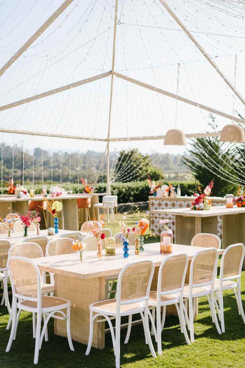 Espaço ao ar livre com mesas e cadeiras de vime e madeira clara, decoradas com arranjos florais coloridos, sob um teto de luzes delicadas