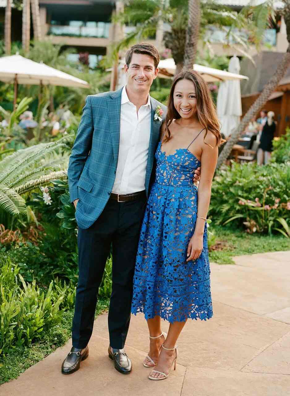 Casal sorridente em um jardim tropical; homem vestindo blazer azul xadrez e calça social e mulher com vestido midi azul rendado.