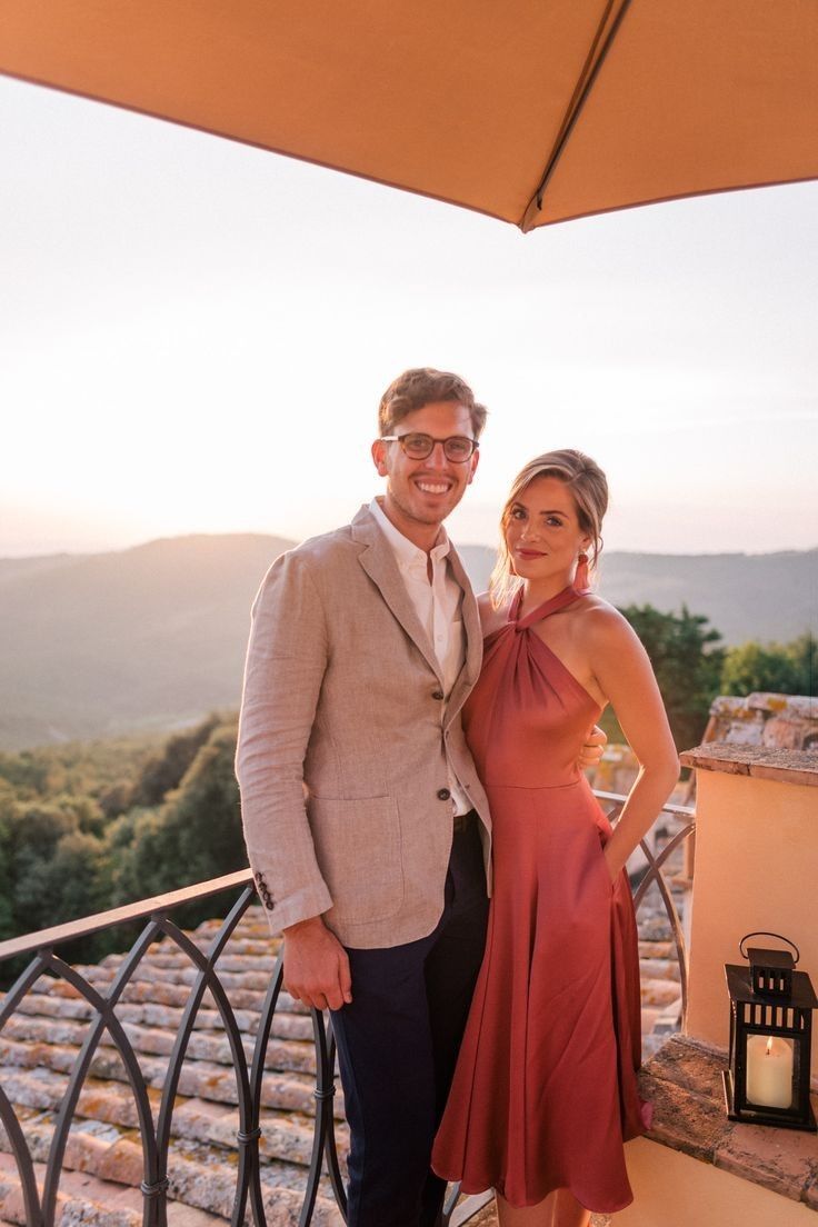 Casal em varanda com vista para o pôr do sol; homem vestindo blazer bege e mulher em vestido vermelho midi com detalhes no pescoço.