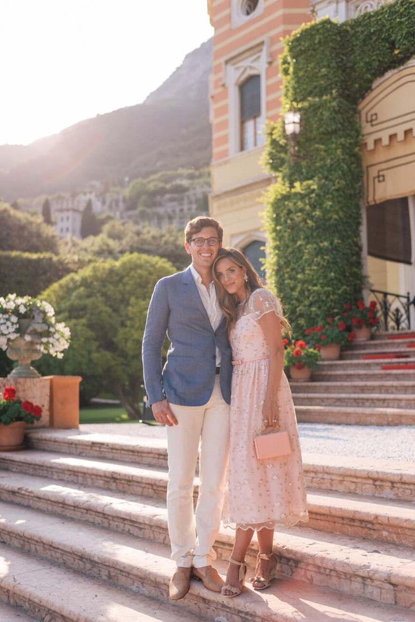 Casal posando em escadaria ao ar livre; homem usando blazer azul e calças claras e mulher com vestido rosa claro e clutch combinando.