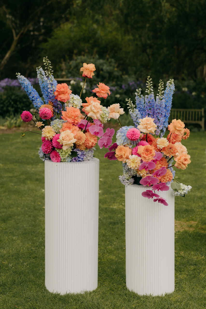 Arranjos florais em tons vibrantes de laranja, rosa e azul, dispostos em vasos brancos altos em um jardim