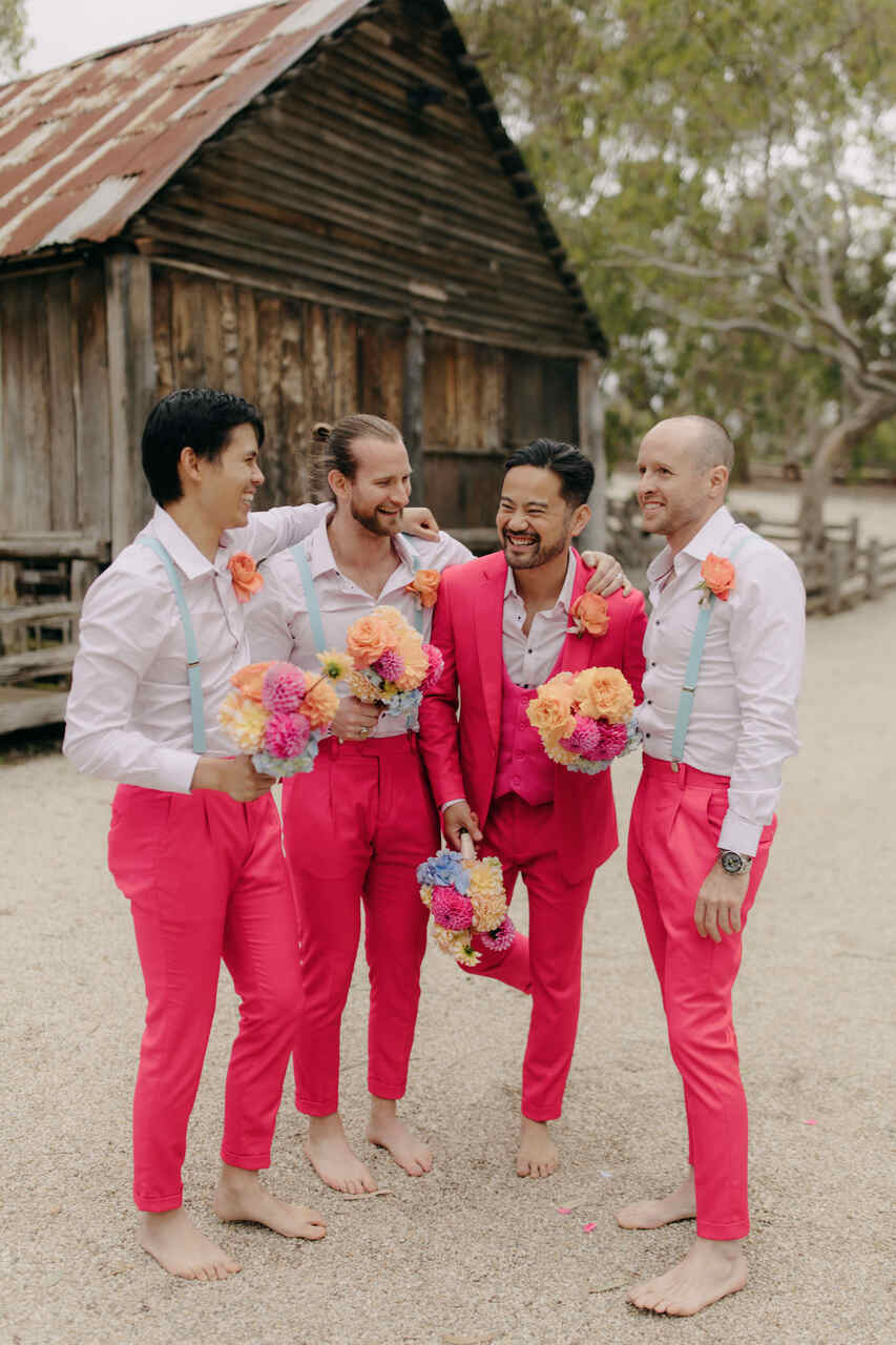 Padrinhos e noivo em trajes cor-de-rosa, combinando com suspensórios e buquês coloridos, posando descalços em frente a um celeiro rústico.