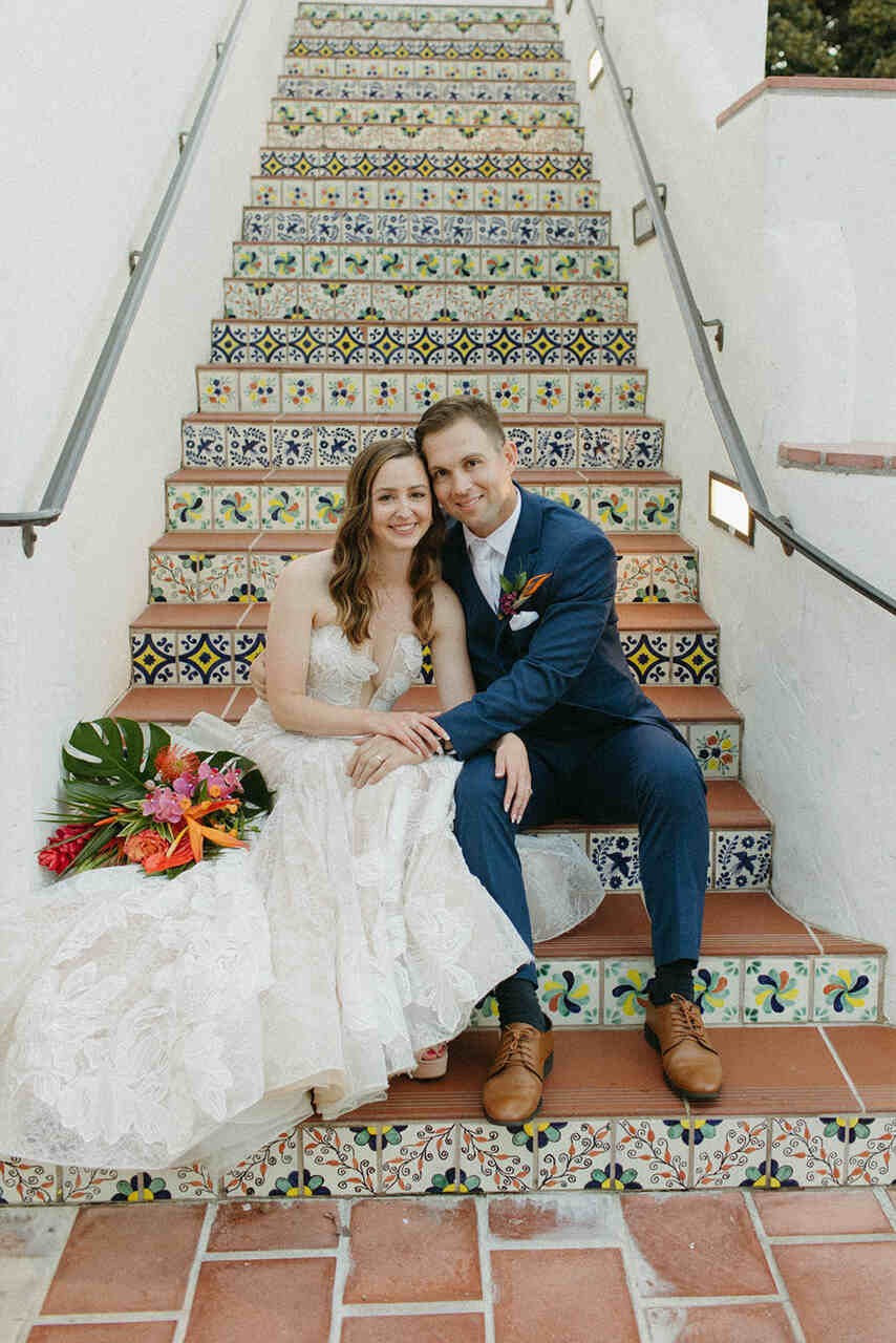 Casal sentado em uma escada com ladrilhos coloridos, segurando um buquê tropical.