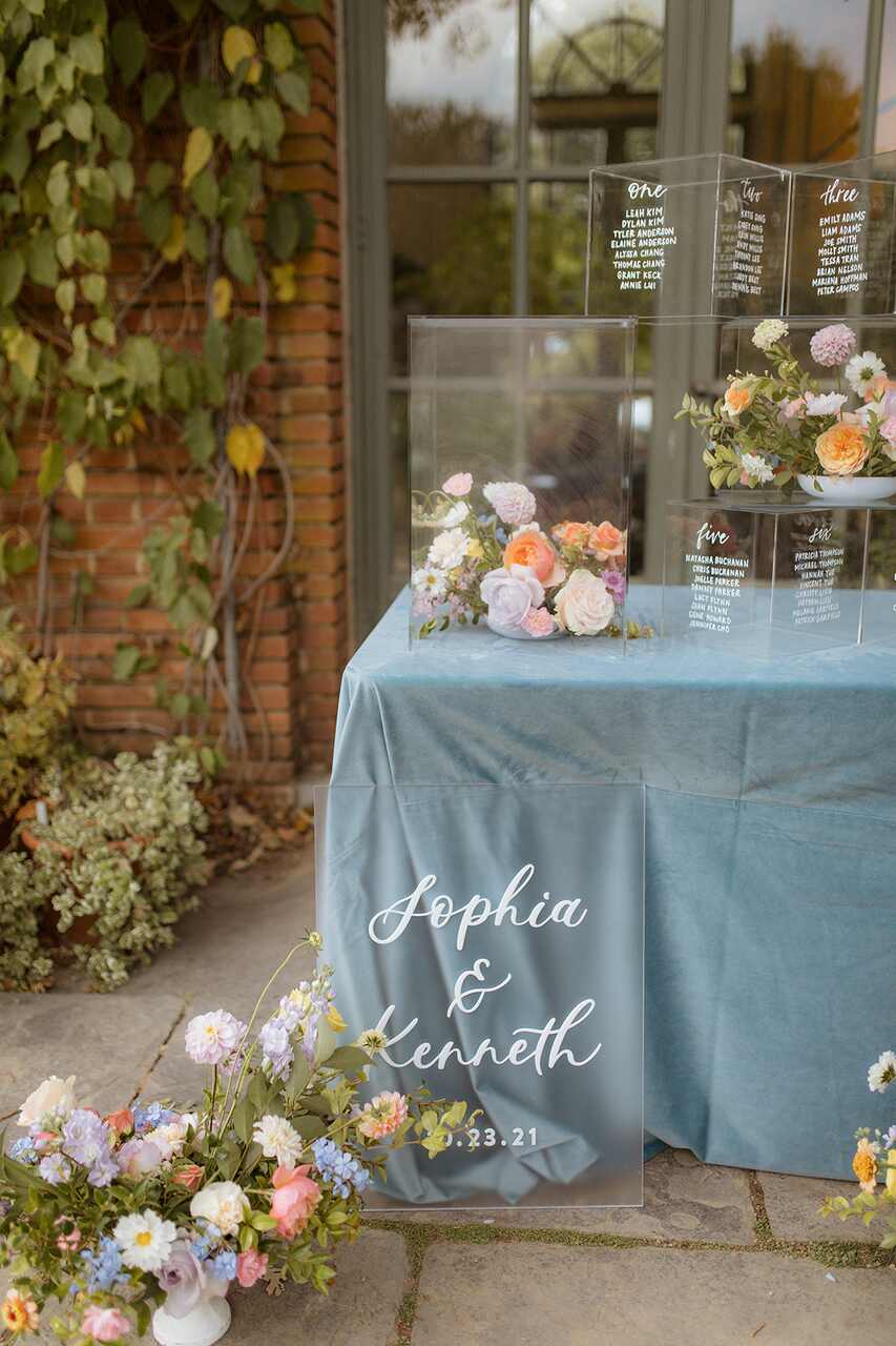 Placa de acrílico elegante com os nomes "Sophia & Kenneth", acompanhada de flores delicadas e uma mesa decorada em tom azul.