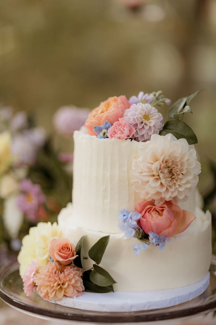 Bolo de dois andares com cobertura texturizada branca, decorado com flores naturais em tons de rosa, pêssego e lilás, posicionado em um ambiente ao ar livre com flores ao fundo.