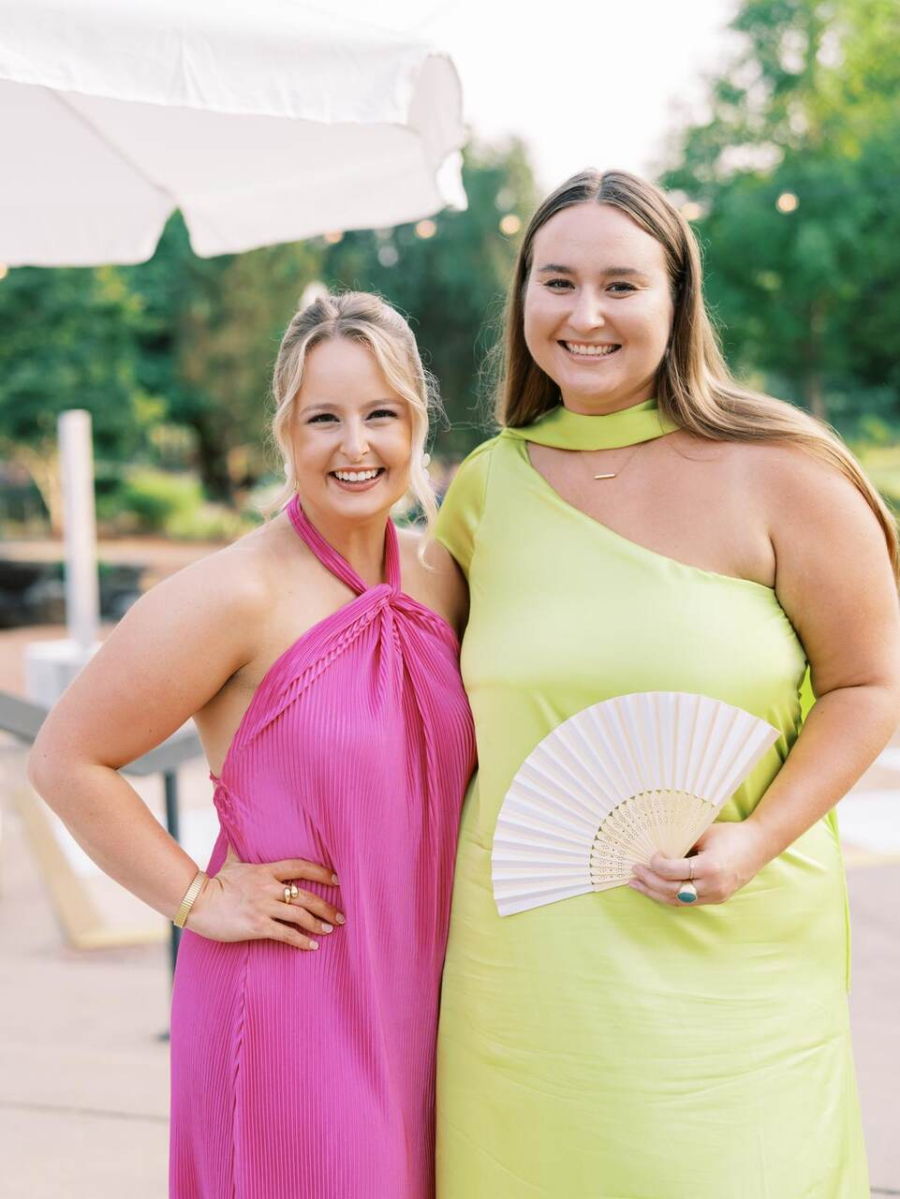 Duas convidadas sorridentes em vestidos elegantes, uma em rosa e outra em verde limão, posando em um ambiente ao ar livre.