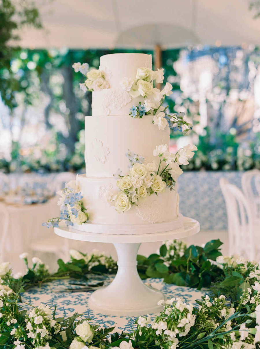 Bolo de casamento clássico de três andares, decorado com flores brancas e delicados detalhes em azul, sobre uma mesa com folhagens e flores brancas ao redor.