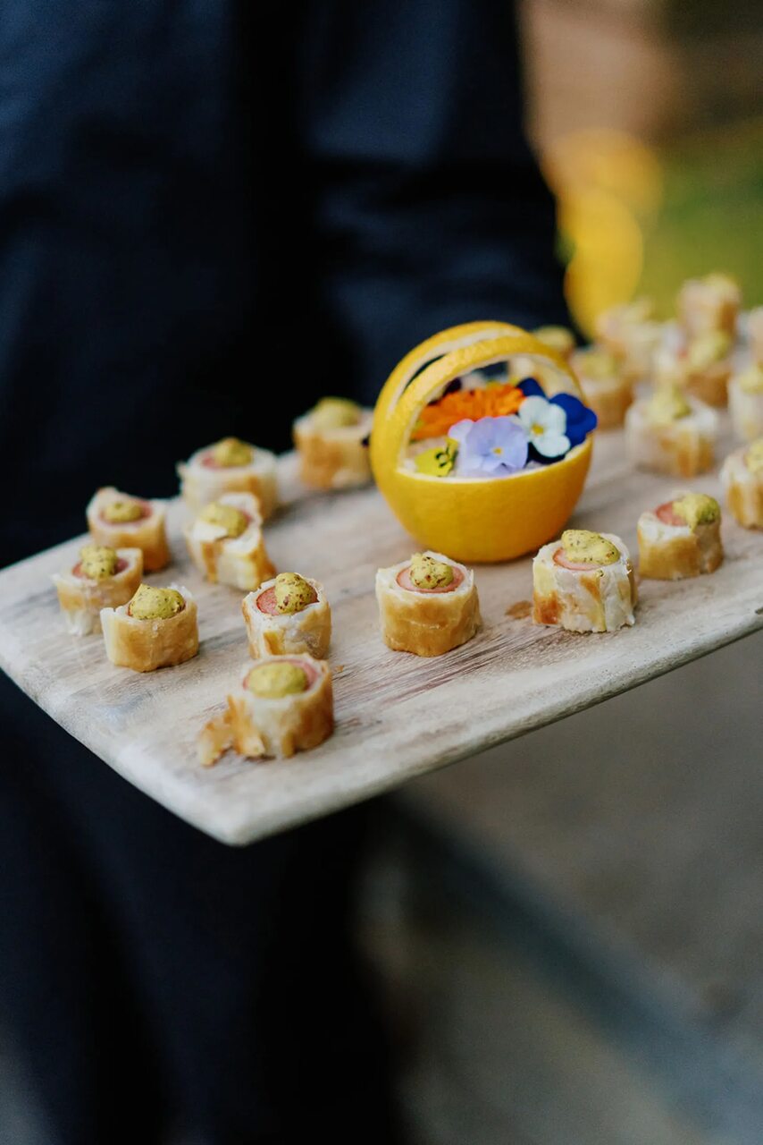 Bandeja de canapés decorada com um pequeno cesto de laranja e flores comestíveis.