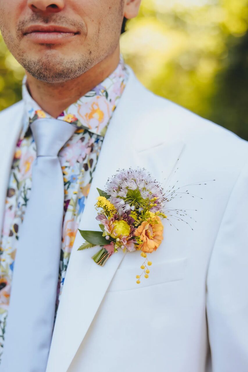 Close de um terno branco com flor delicada e colorida na lapela, complementando uma camisa estampada floral.