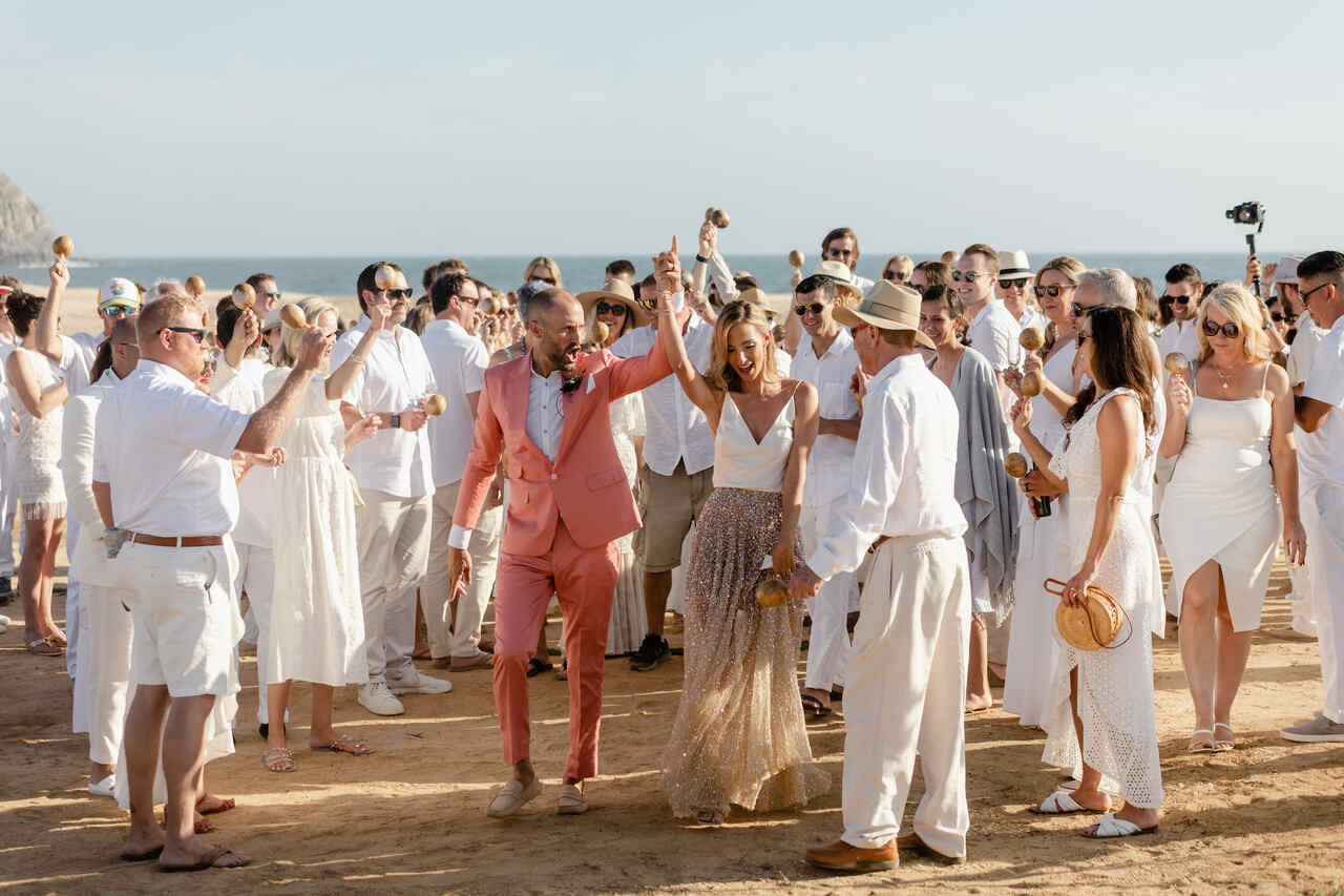 Casamento na praia com noivo usando terno rosa e noiva em vestido dourado, cercados por convidados em roupas brancas.