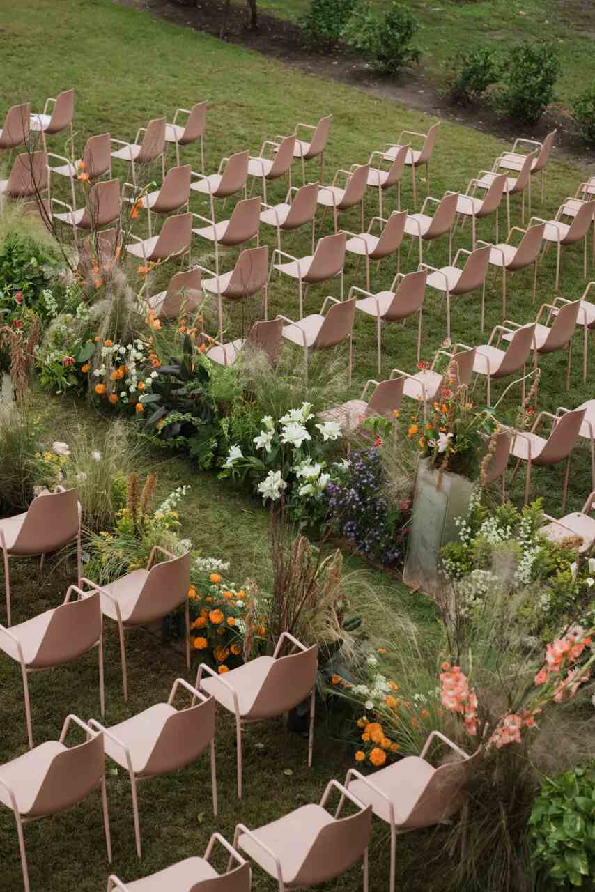 Arranjo de cadeiras em um jardim ao ar livre, dispostas em fileiras simétricas, cercadas por uma decoração exuberante de flores em tons vibrantes, como laranja e branco, criando um corredor central florido.