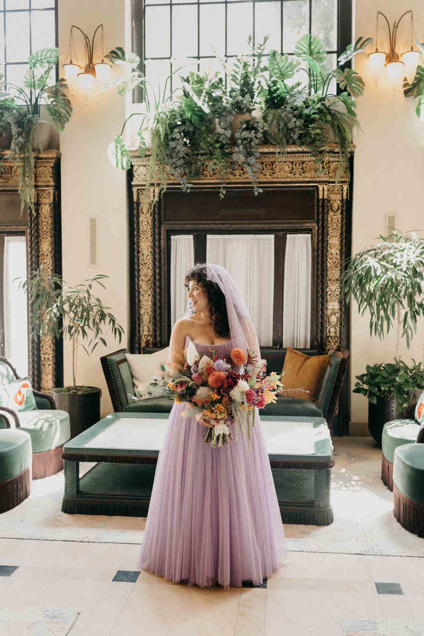 Noiva em um vestido de tule lilás, segurando um buquê vibrante, posando em um ambiente interno com decoração luxuosa e plantas exuberantes.