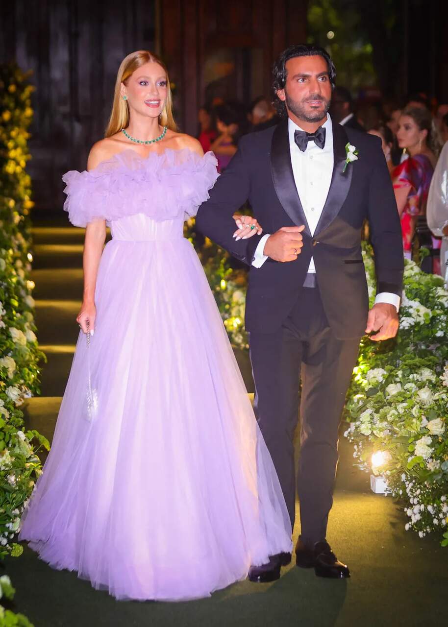 Casal em evento noturno; mulher vestindo um vestido lilás volumoso com detalhes em tule e homem usando smoking preto com gravata borboleta.