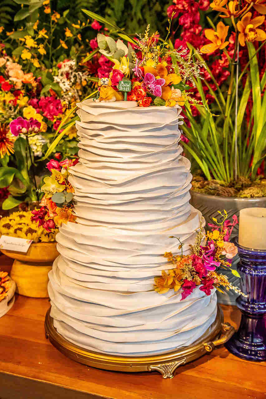 Bolo de casamento de três camadas com detalhes texturizados e flores tropicais em tons vibrantes no topo.