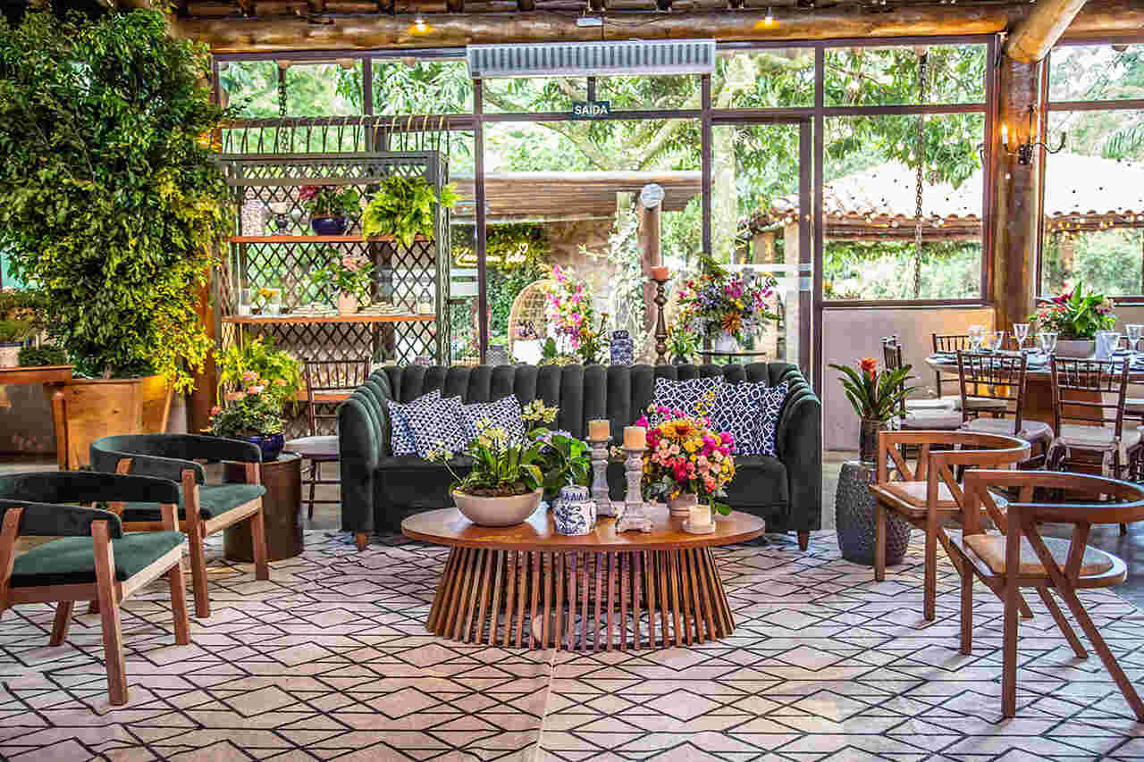 Lounge de casamento com decoração vibrante e aconchegante, composto por sofá verde, poltronas de madeira, almofadas estampadas e arranjos florais coloridos.