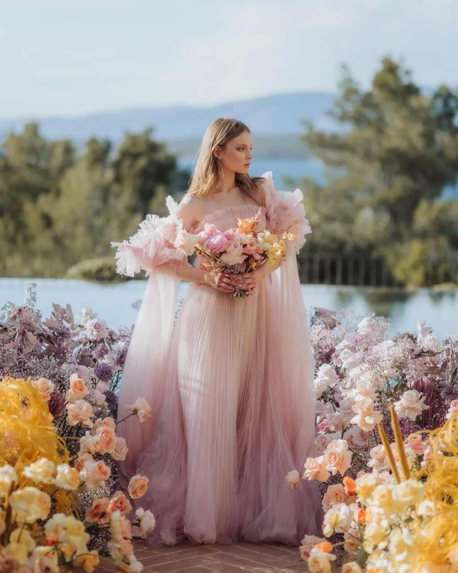 Noiva posando em meio a um corredor repleto de flores vibrantes em tons de rosa, amarelo e lilás, com vista para o mar ao fundo. Seu vestido leve em tons de rosa claro