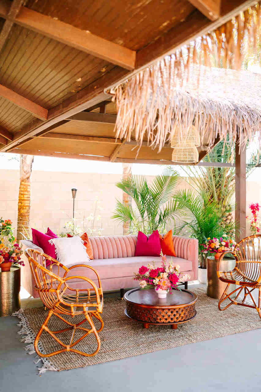 Área lounge para casamento tropical com sofá rosa, cadeiras de vime, flores coloridas e detalhes rústicos