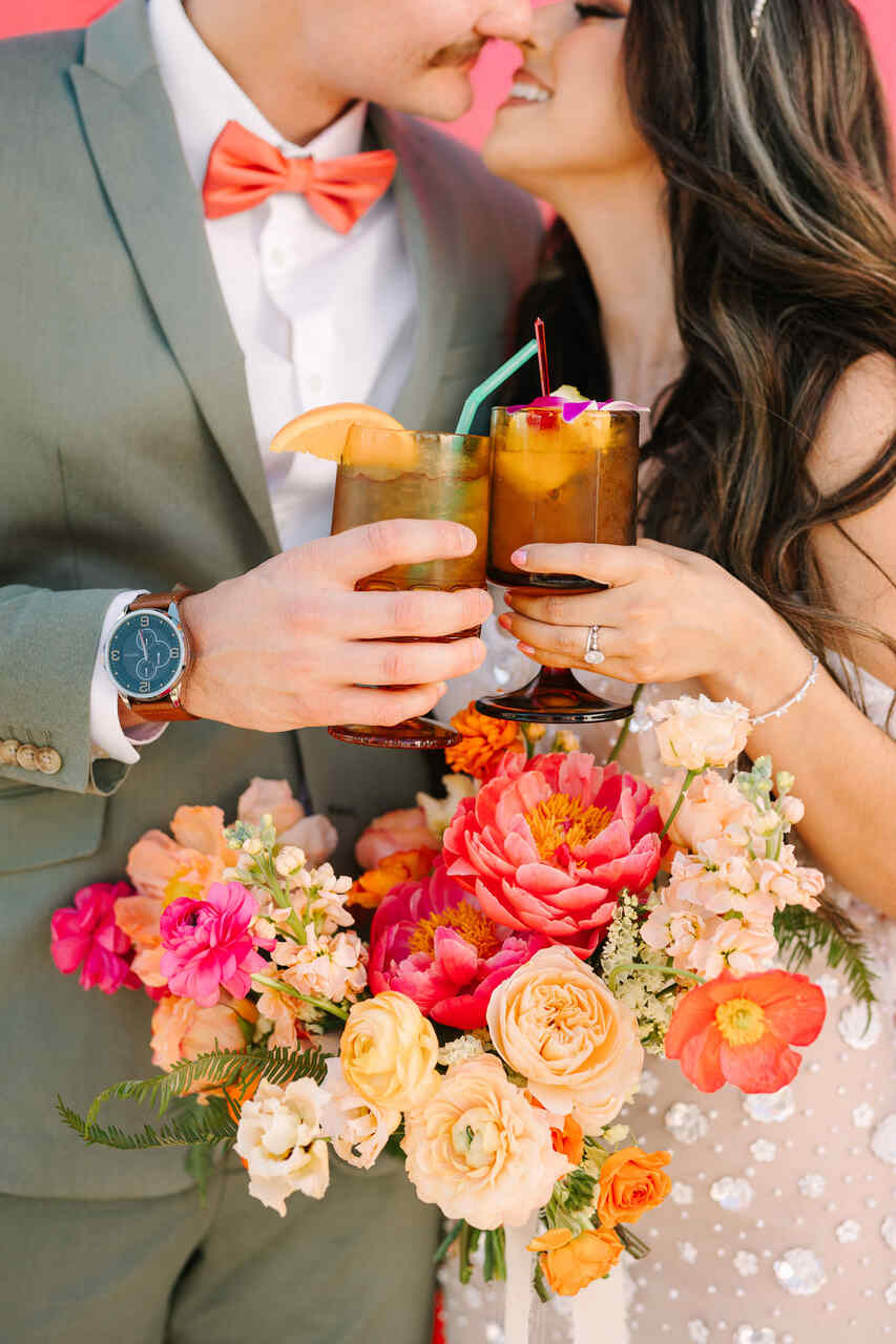  Casal brindando com coquetéis em mãos, cercado por flores vibrantes e um fundo colorido.