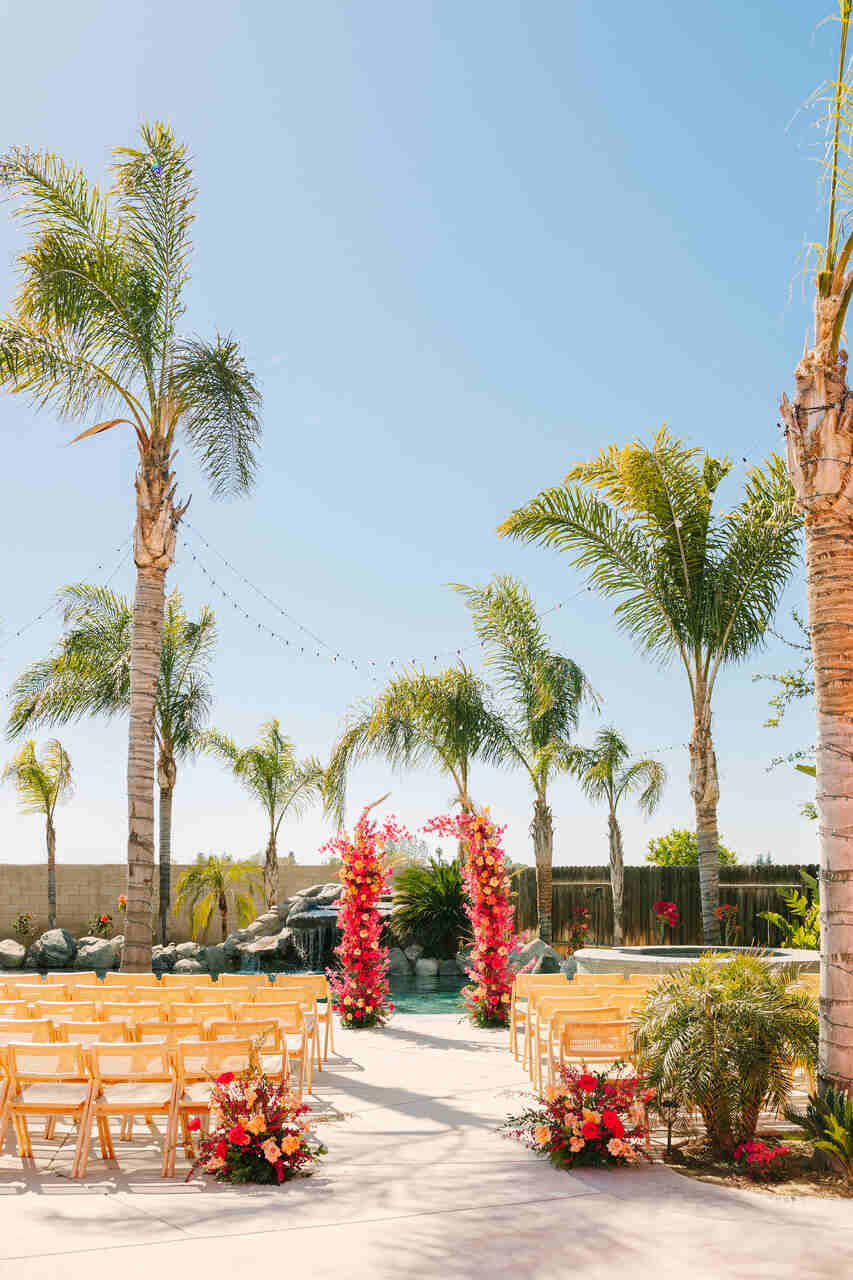 Cerimônia de casamento ao ar livre com cadeiras dispostas em fileiras, arco floral vibrante em tons de rosa e laranja, palmeiras ao redor e céu azul ao fundo, criando um cenário tropical.