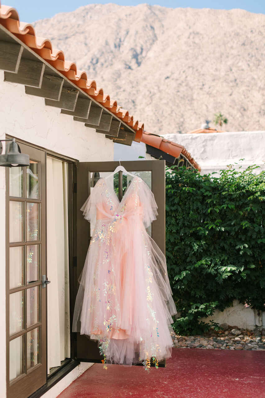 Vestido de noiva com tule rosa e detalhes holográficos pendurado em uma porta aberta, com um cenário de montanhas ao fundo e um ambiente ensolarado.