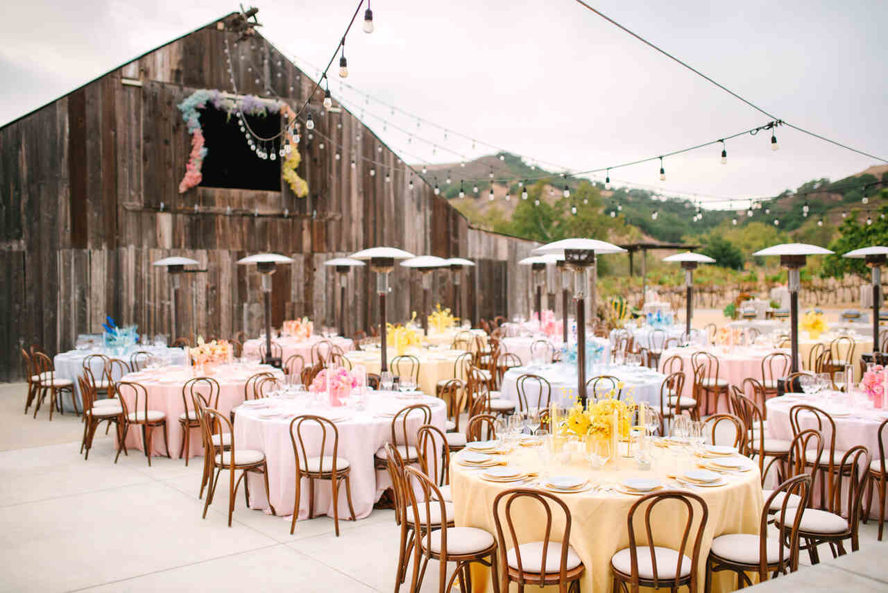 Cenário ao ar livre com mesas redondas decoradas em tons pastel, cercadas por cadeiras de madeira, com um celeiro rústico ao fundo e luzes penduradas criando um clima aconchegante.