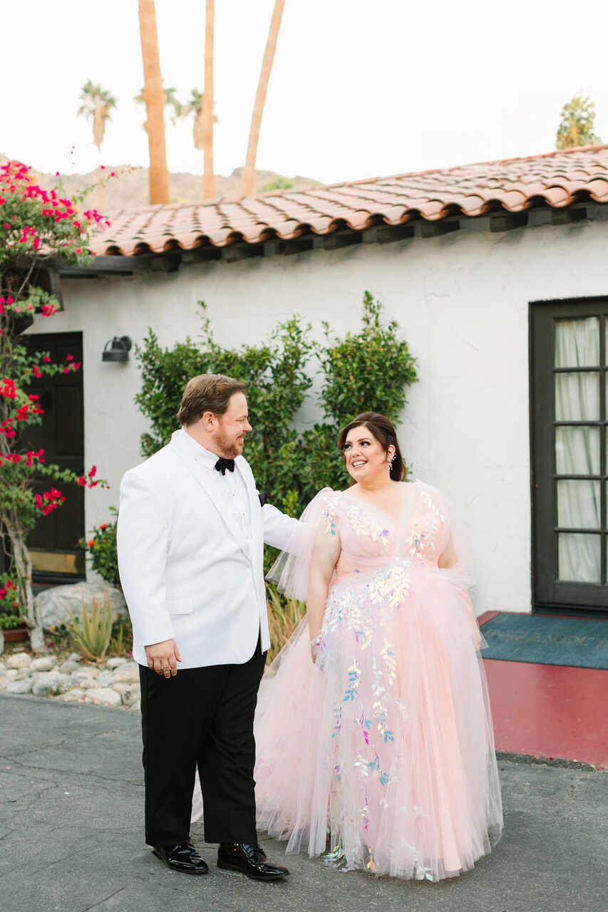 Casal sorridente ao ar livre, com a noiva em um vestido de tule rosa com brilhos holográficos e o noivo em um elegante terno branco.