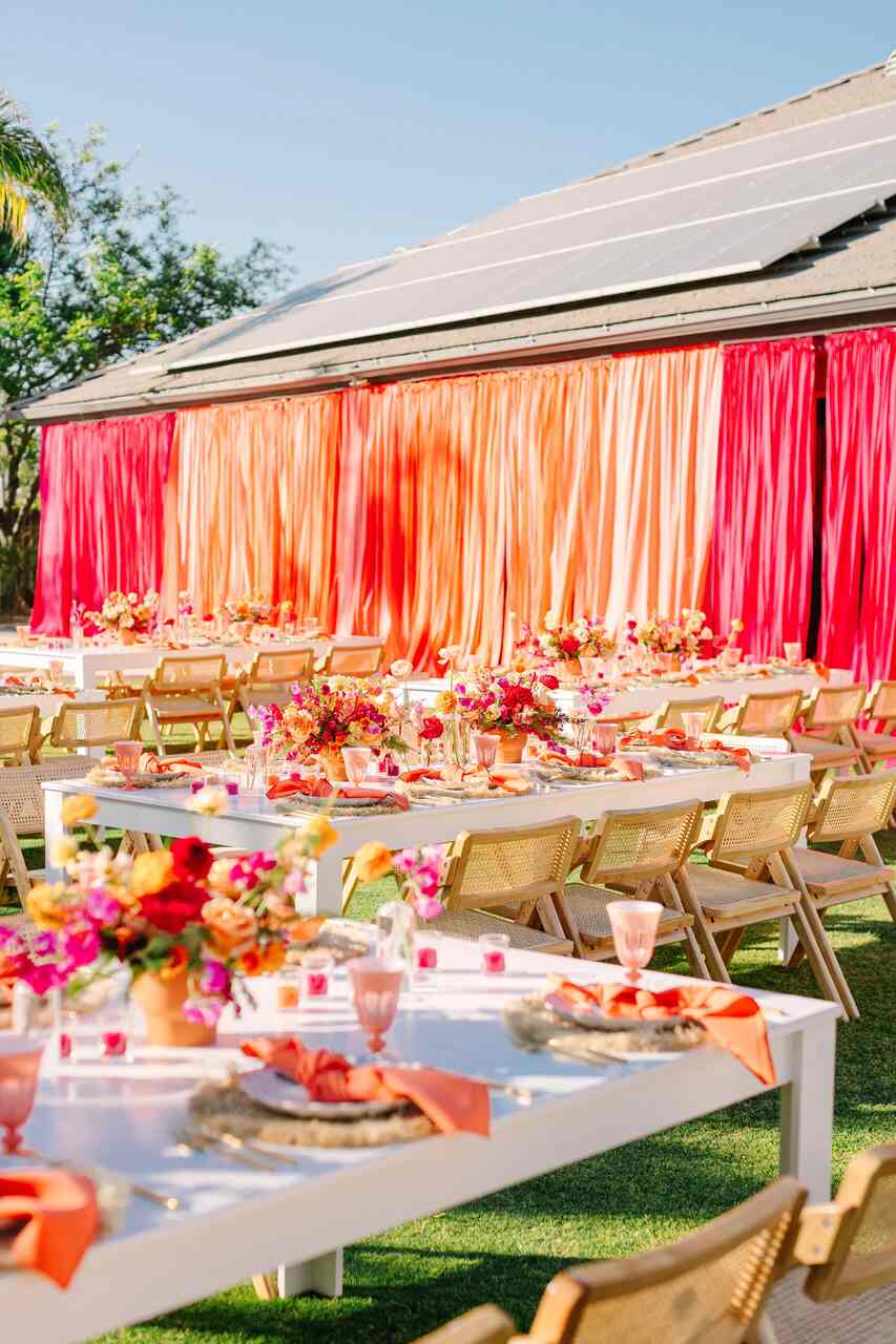 Mesas retangulares brancas decoradas com arranjos florais e cortinas coloridas em tons quentes ao fundo, em uma festa ao ar livre