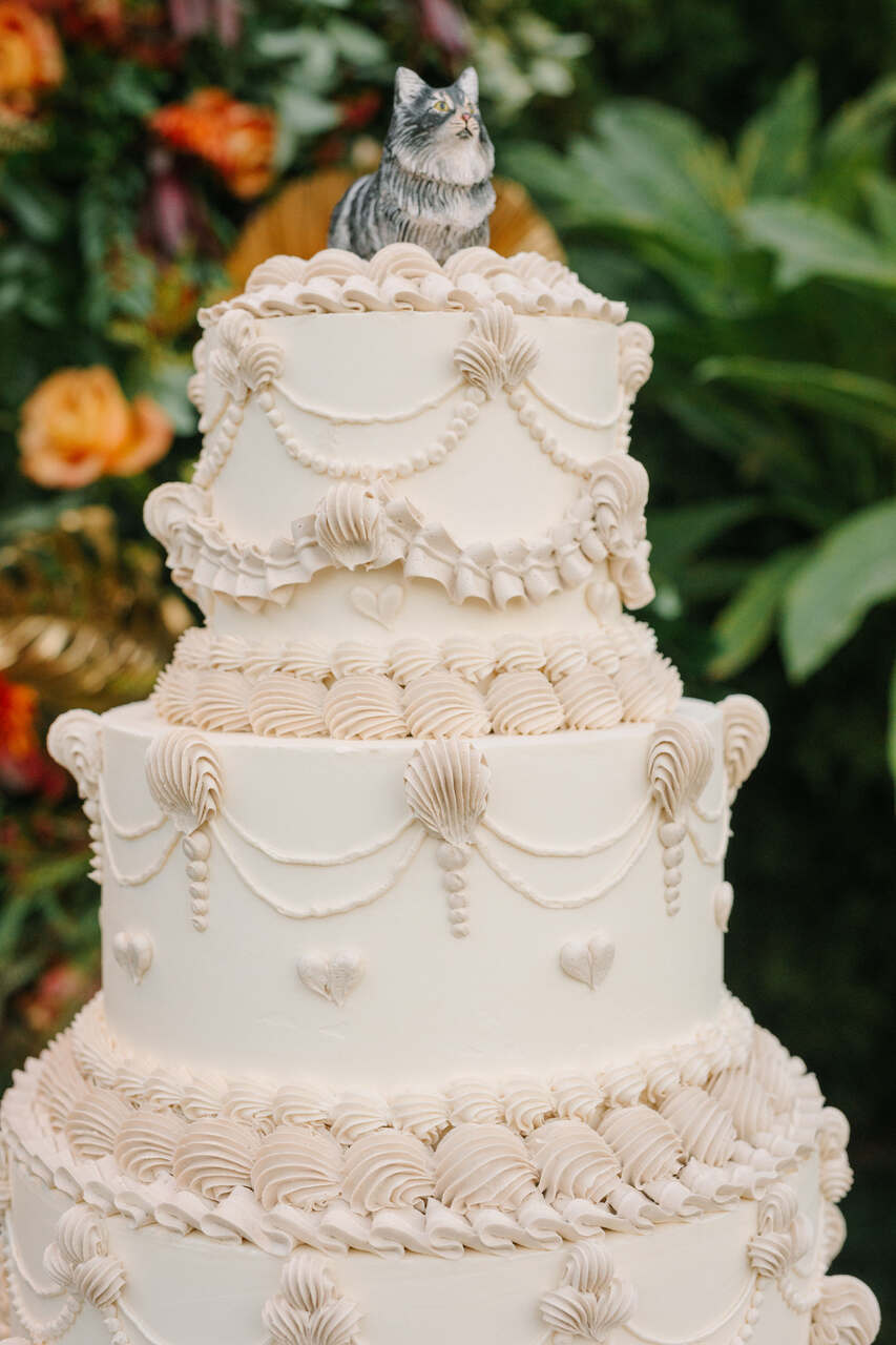 Bolo branco de três andares com detalhes decorativos em glacê que simulam babados e pérolas, finalizado com um topo em formato de gato e detalhes delicados de conchas.