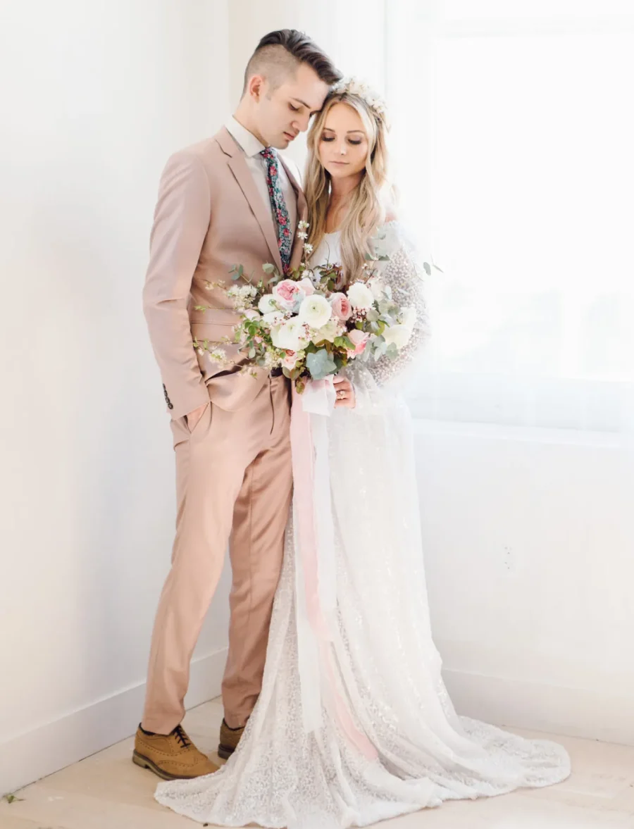 Casal em traje de casamento, o noivo com terno rosa claro e a noiva em vestido branco rendado, ambos em um ambiente minimalista.