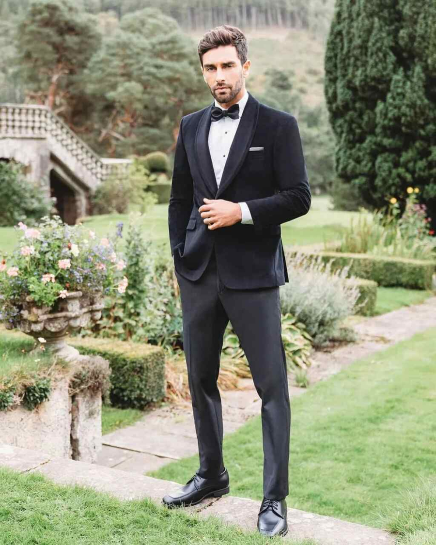 Homem em jardim elegante, usando smoking preto com gravata borboleta, posicionado em frente a uma paisagem clássica.