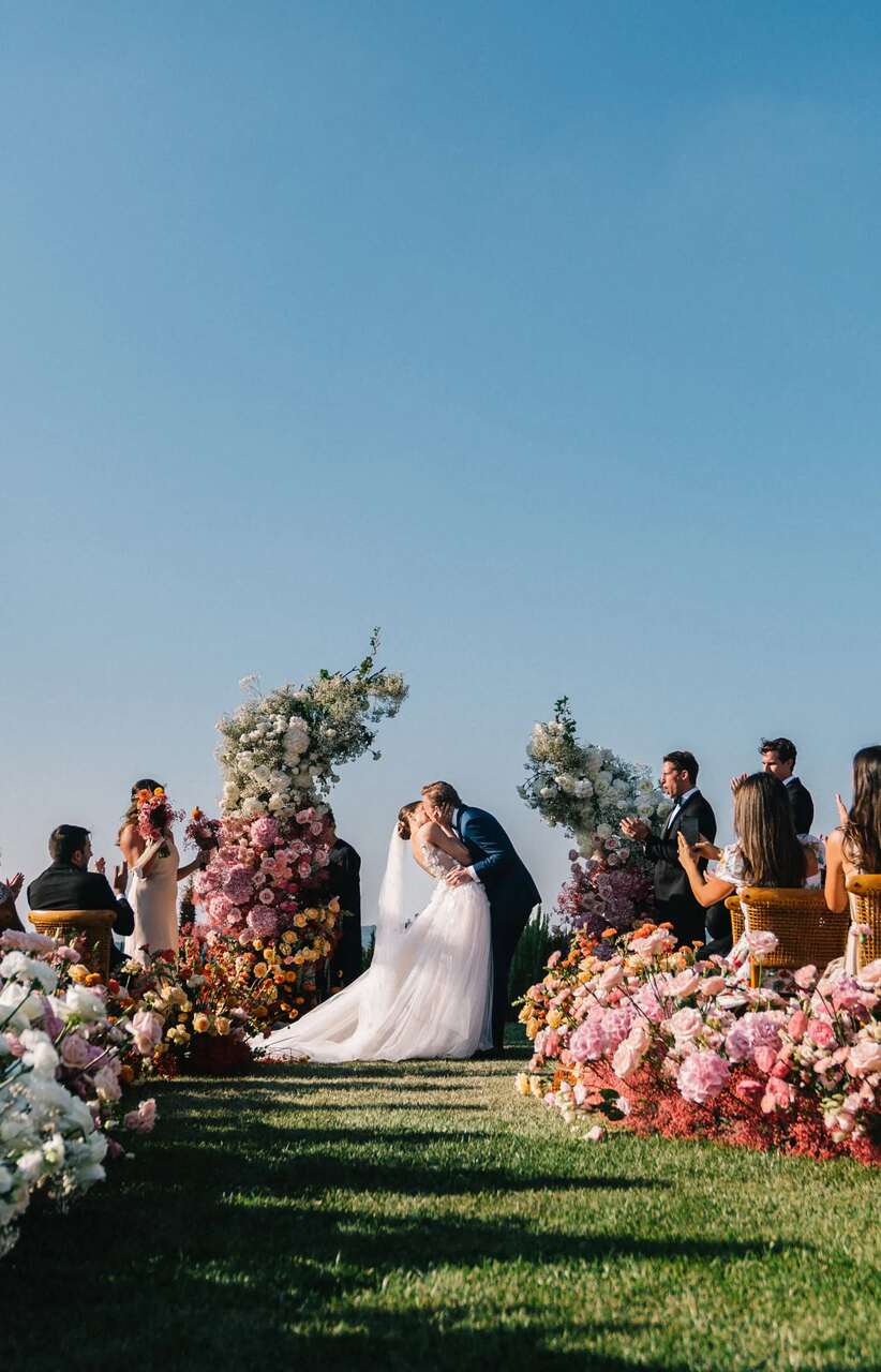 Casal se beijando ao final da cerimônia de casamento ao ar livre, com um corredor decorado por flores em tons pastel e céu azul ao fundo, enquanto convidados aplaudem.