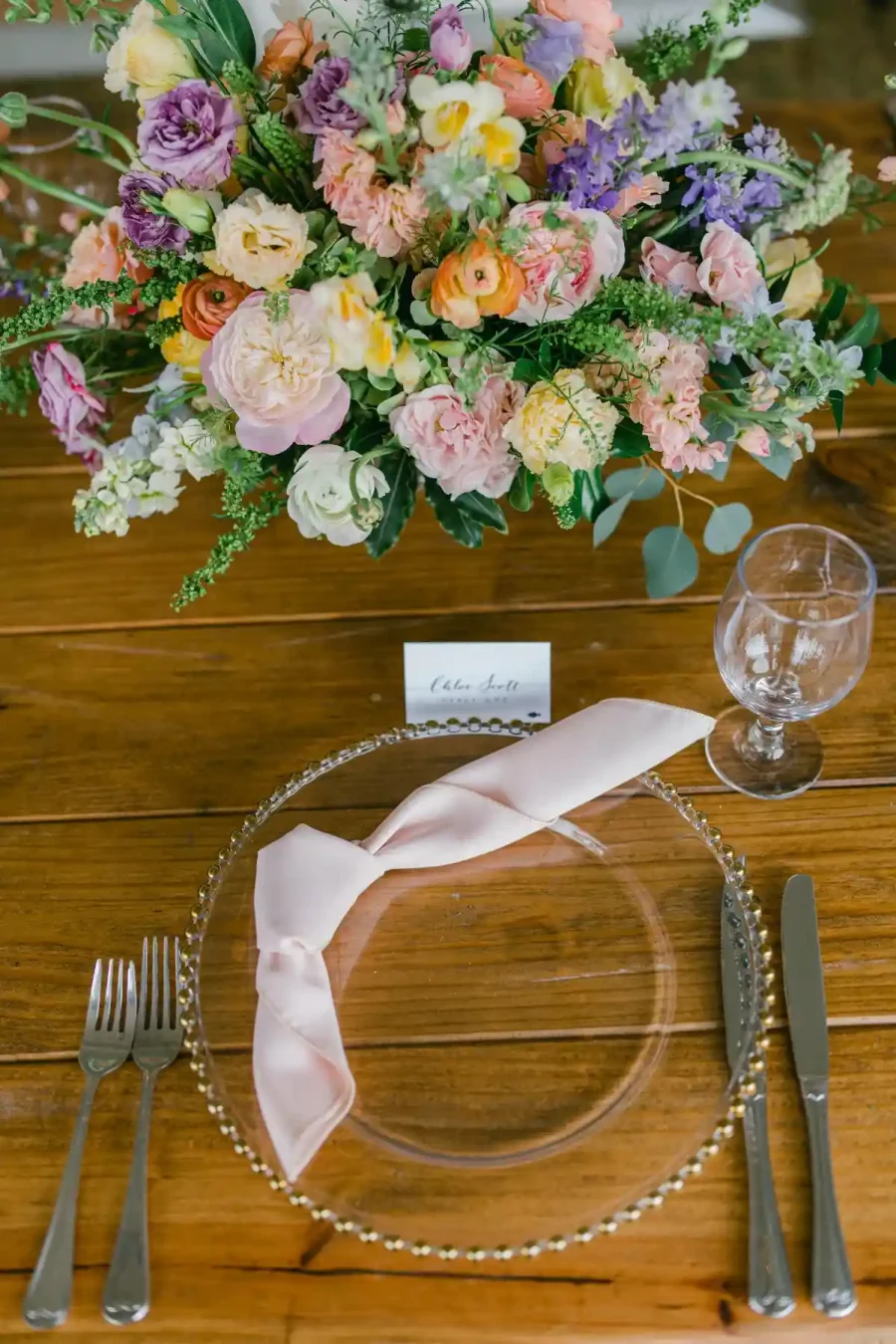 Configuração de mesa com um prato transparente em um sousplat de contas douradas, decorado com um guardanapo rosa claro dobrado elegantemente. Há talheres de prata e uma taça de vidro ao lado. Um arranjo floral exuberante com flores de cores vivas está no centro.