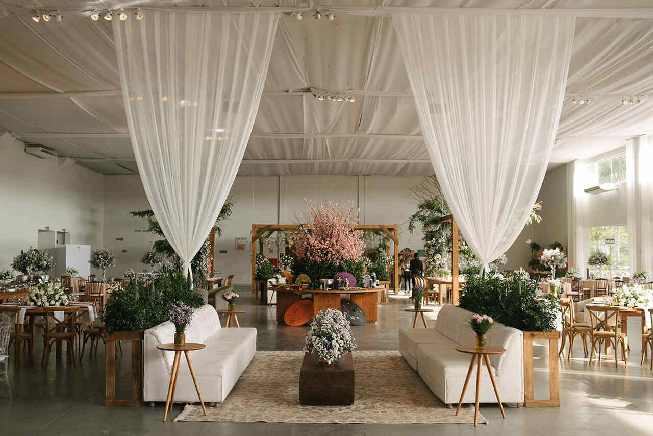 Lounge em casamento com decoração clássica e romântica, com sofás brancos, mesas de apoio de madeira, cortinas leves e arranjos florais delicados.