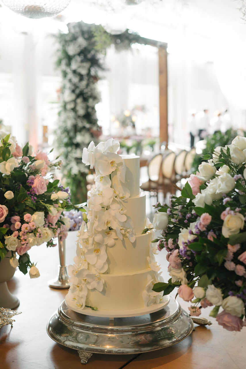 Um elegante bolo de quatro andares com detalhes florais coloridos que o envolvem, acompanhado de arranjos florais em tons pastel sobre uma mesa de madeira.