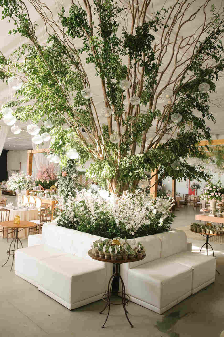 Lounge centralizado ao redor de uma grande árvore decorada com flores brancas e luzes pendentes, combinando sofás brancos e mesas delicadas